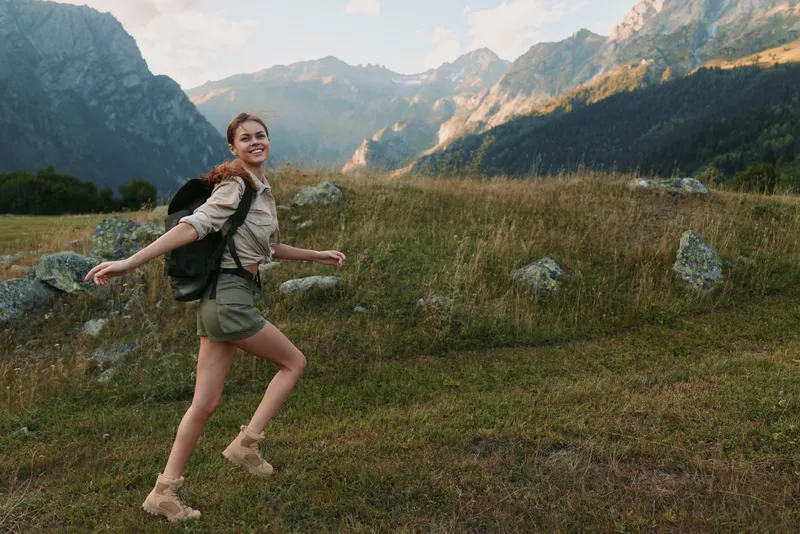 Young woman hiking outdoors in a grassy meadow with rugged mountains in the background, backpack on her shoulders, smiling as she moves forward under bright daylight.