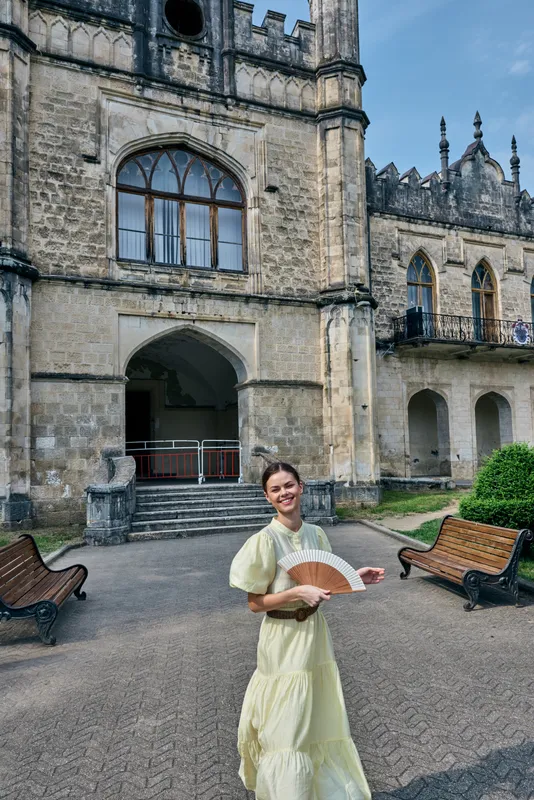 A woman in a pale dress stands in a historic castle courtyard, holding a fan, smiling amid ancient architecture and an outdoor travel scene.