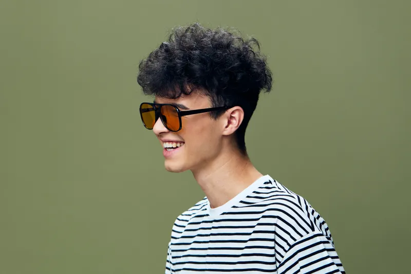 Smiling young man with curly hair wearing orange sunglasses and a striped shirt against a green background, expressing happiness and casual style.