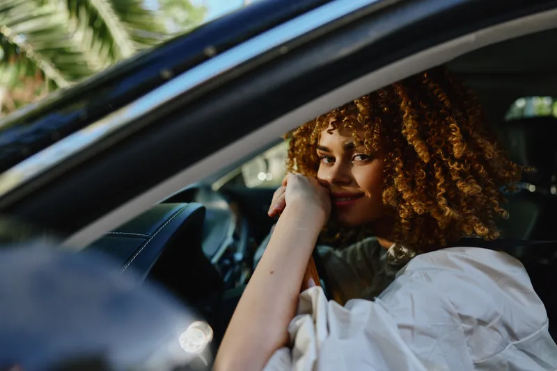 Smiling woman with curly hair sitting inside a car, resting her arms on the steering wheel, illuminated by natural light and surrounded by greenery outside the window.