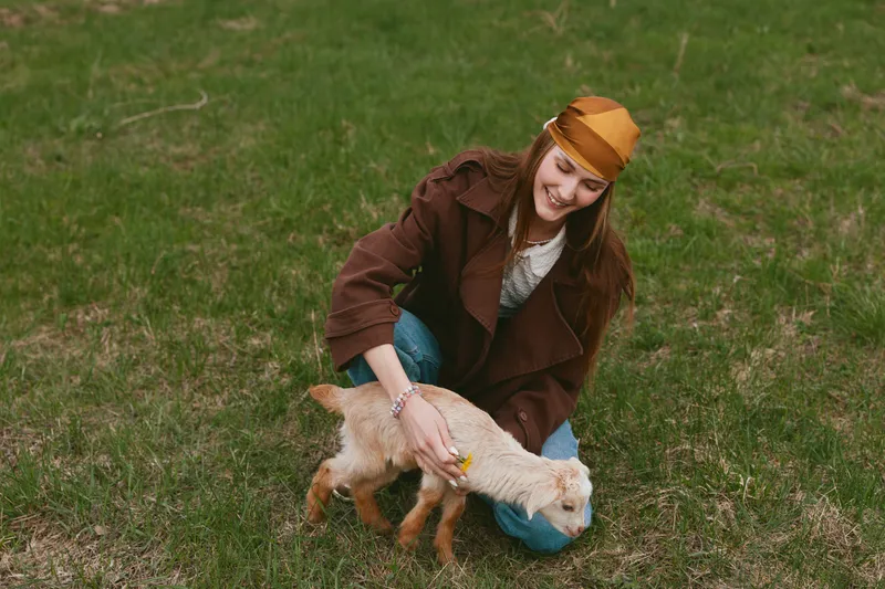 Cheerful woman outdoors, kneeling beside a playful goat kid, gentle touch, sunlit grass, relaxed posture, casual clothing, warm tones, candid lifestyle moment with animals