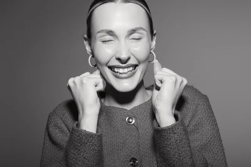 Happy smiling white woman with fair skin in grey sweater closes eyes and clenches fists in joy isolated on studio background.