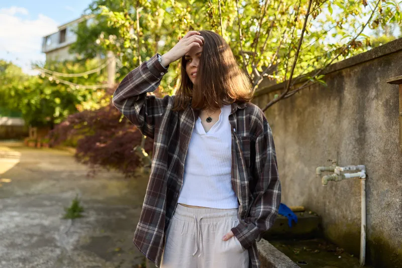 A casual woman outdoors wearing a plaid shirt and white tee, adjusting her hair beside a sunlit wall. A lifestyle portrait capturing everyday calm and natural movement