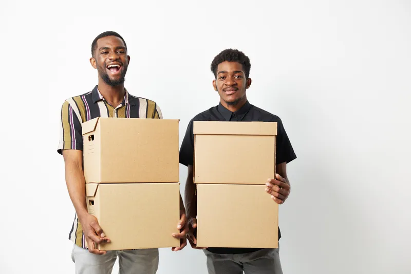 Two smiling African American men holding cardboard boxes, demonstrating teamwork and collaboration, set against a clean, minimalistic background.