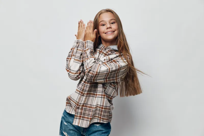 Girl child with long hair smiling and wearing plaid shirt and jeans posing with hands together on gray background. Happy kid, casual outfit, cheerful and confident expression.