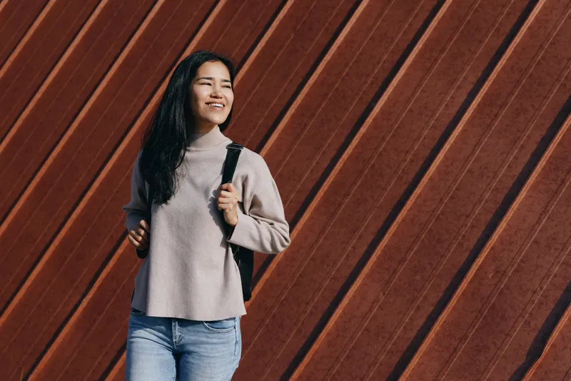 Woman with backpack standing against a diagonal wooden wall, wearing a cozy sweater and jeans, smiling as she heads to travel or study in a casual street scene