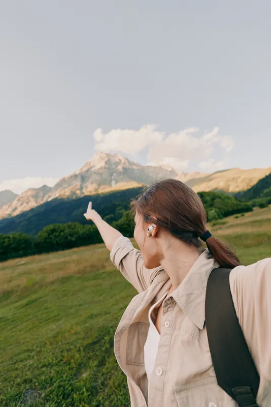 Woman in outdoor lifestyle with backpack stands in a meadow, pointing toward distant mountains, embracing nature and scenic landscape under a clear sky, suggesting adventure