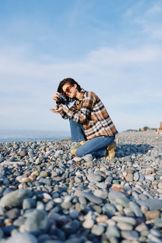 Photographer on a pebble beach kneels to shoot with camera, casual plaid shirt and jeans, blue sky and calm sea form a vast backdrop for coastal photography