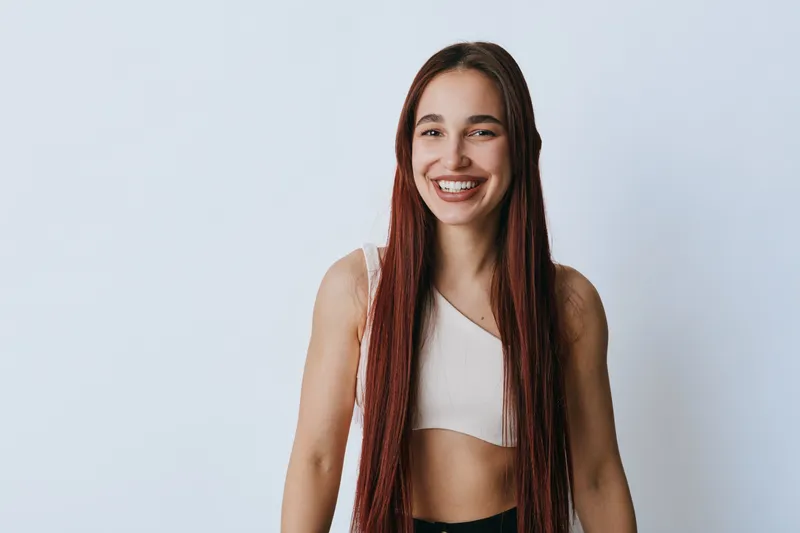 Young woman with long braided hair smiling and standing against white background. Casual clothing and natural expression in studio portrait with bright lighting and relaxed mood.
