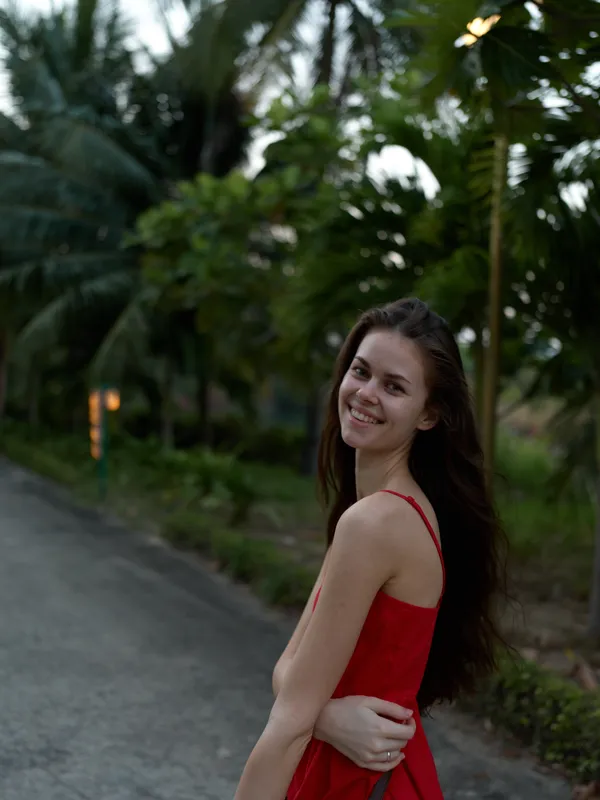 smiling young woman in red dress standing outdoors on pathway, natural light with green trees and plants in background, casual summer portrait