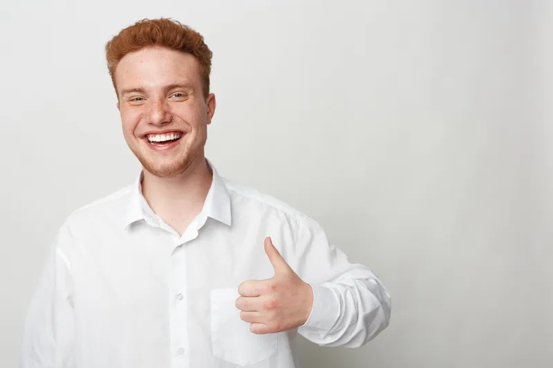Smiling young man with red hair and freckles wearing white shirt shows thumbs up gesture. Positive emotion, casual style, isolated on light background. Concept of approval and happiness.