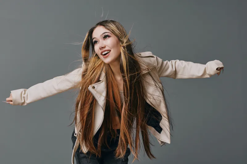 Young woman with long hair wearing light jacket and black pants posing with arms extended on gray background. Casual style, hairstyle and confident facial expression in studio photo.