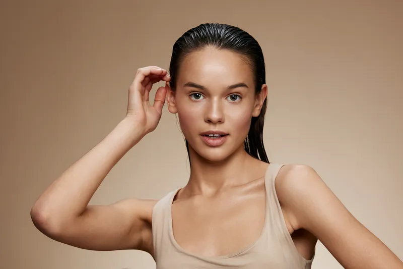 Young model with wet hair and natural makeup posing against a neutral background, showcasing beauty, confidence, and skincare