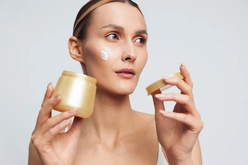 Young woman applying cream on face holding golden jar and lid, clear skin, natural beauty, skincare routine, neutral background, focused expression, healthy lifestyle
