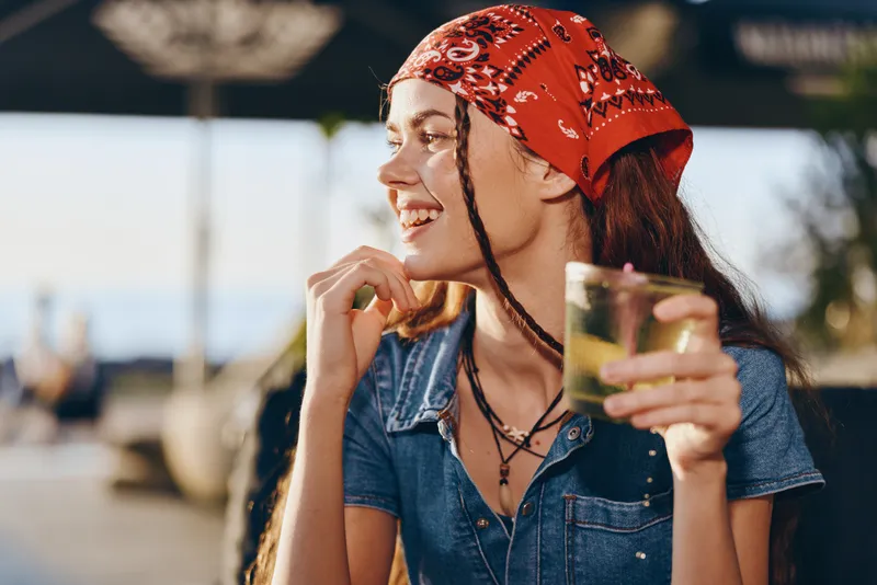 smiling woman with dreadlocks wearing red bandana holding drink outdoors in sunny casual denim outfit enjoying summer atmosphere and relaxed mood