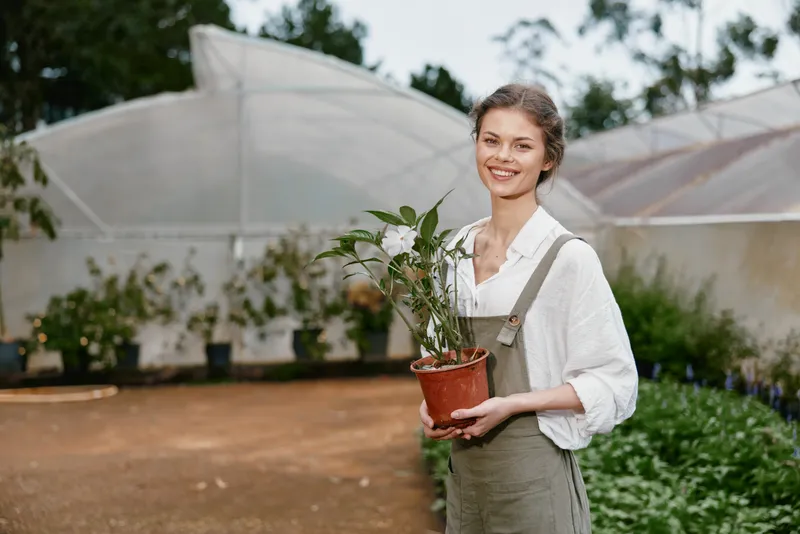 Smiling young woman in overalls holding a potted plant in a greenhouse, surrounded by greenery and natural light, showcasing sustainable gardening and happiness