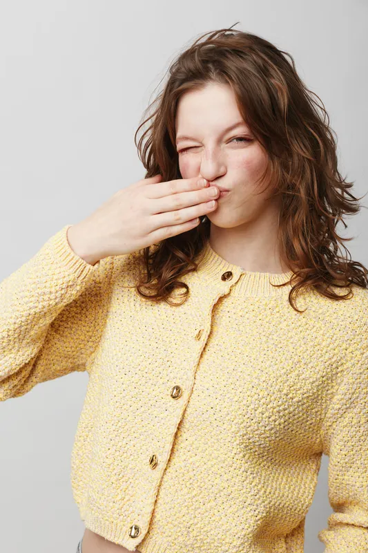 Young woman in yellow knitted cardigan covers mouth with hand, winking and smiling. Studio portrait on light background. Concept of playful gesture, happiness, and casual fashion.