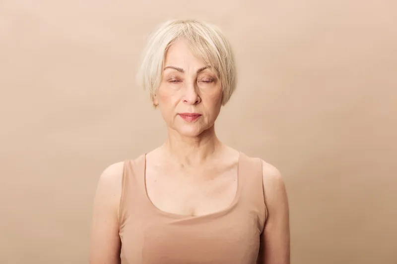Calm mature woman with short grey hair and closed eyes stands against beige background wearing sleeveless top. Concept of mindfulness, relaxation, aging, and natural beauty in studio lighting.