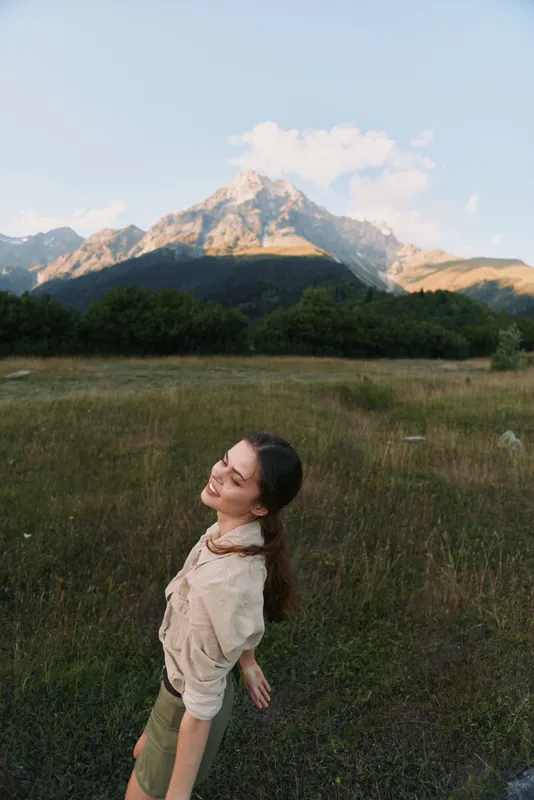 Young woman wearing a beige sweater and khaki shorts stands in a grassy meadow with a mountain backdrop, smiling warmly at the camera under a clear blue sky on a sunny day.