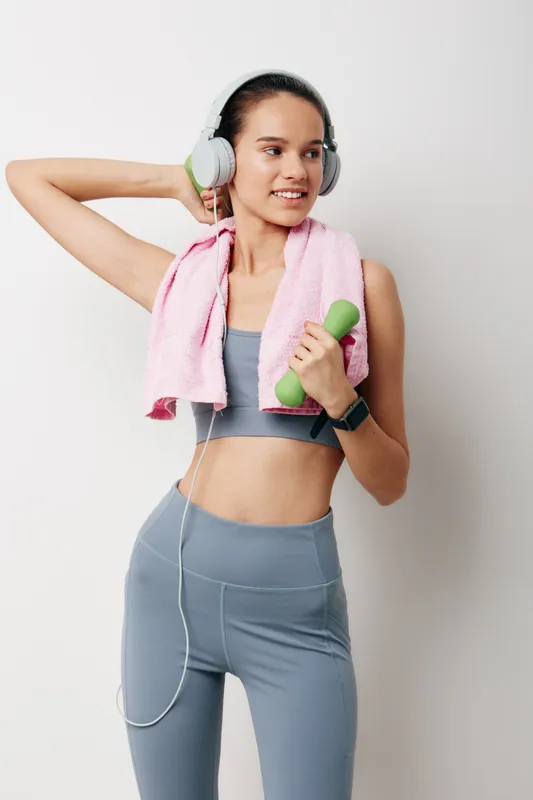 Fitness oriented woman in athletic wear poses with a dumbbell and pink towel, wearing headphones in a clean studio, ready for a focused workout and strong performance.