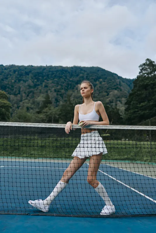 Athletic young woman in white skort and crop top poses confidently on a tennis court, showcasing a sporty vibe and vibrant outdoor background