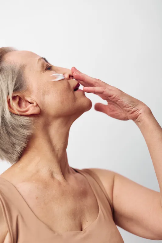 Mature woman with short grey hair applies face cream on cheek in beige top. Close-up, skincare routine concept focused on aging skin care, beauty, and self-care with soft natural lighting.