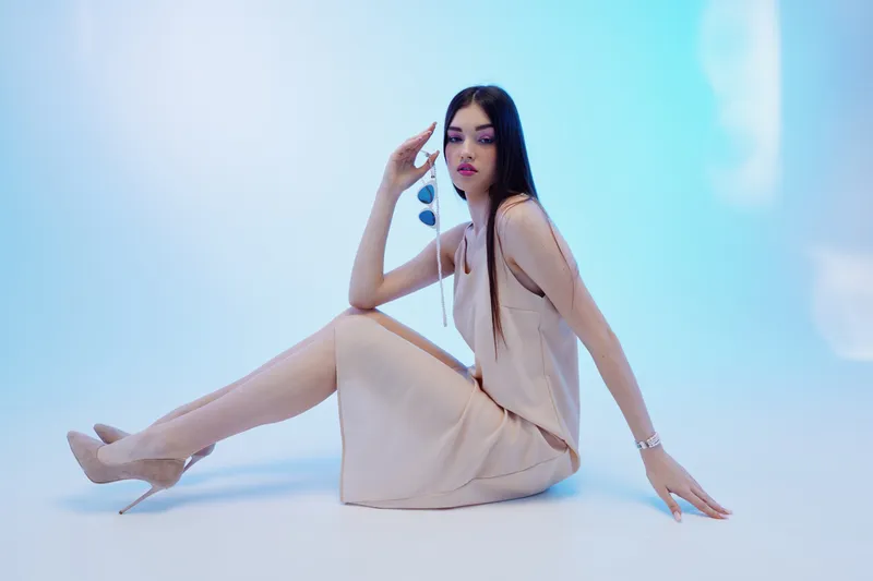 Elegant woman in a beige dress and high heels sits on the floor in a studio with soft blue lighting. Concept of beauty, fashion, and modern style.