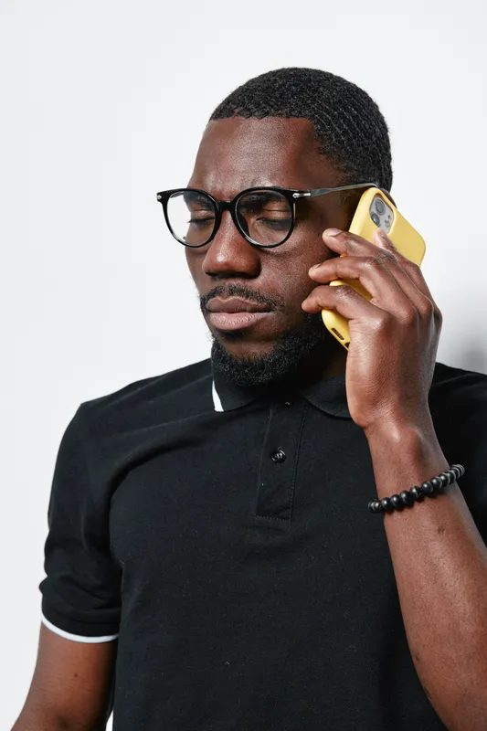 African American man with dark skin wearing glasses and black polo shirt talking on yellow smartphone isolated on white background. Serious expression and casual style.