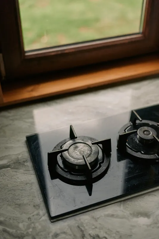 Stove with two gas burners sits on a stone countertop near a window, showcasing a clean modern kitchen setup and a reflective glass surface.