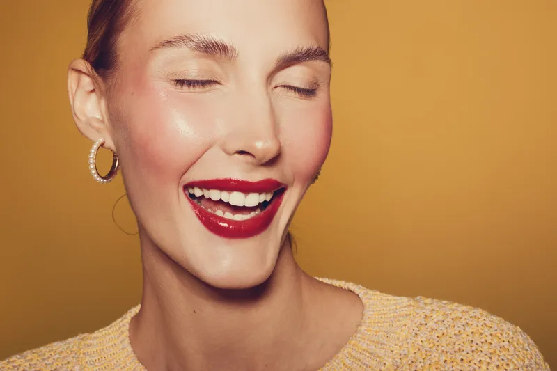 Close-up of happy woman with red lipstick smiling with closed eyes on yellow background. Concept of beauty, makeup, joy, and positive emotions in studio lighting.