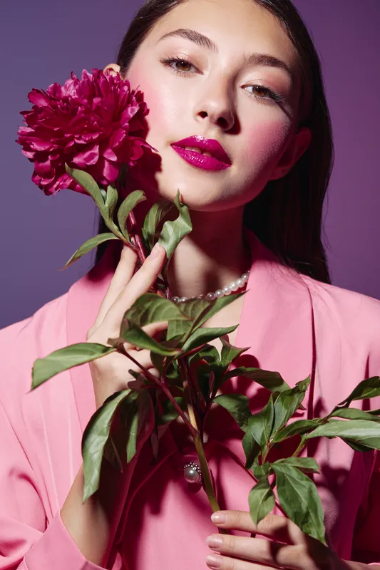 Young woman with trendy glossy fuchsia lips and pink blush holds a fresh red peony against a purple background. Creative beauty fashion concept emphasizes vibrant colors.