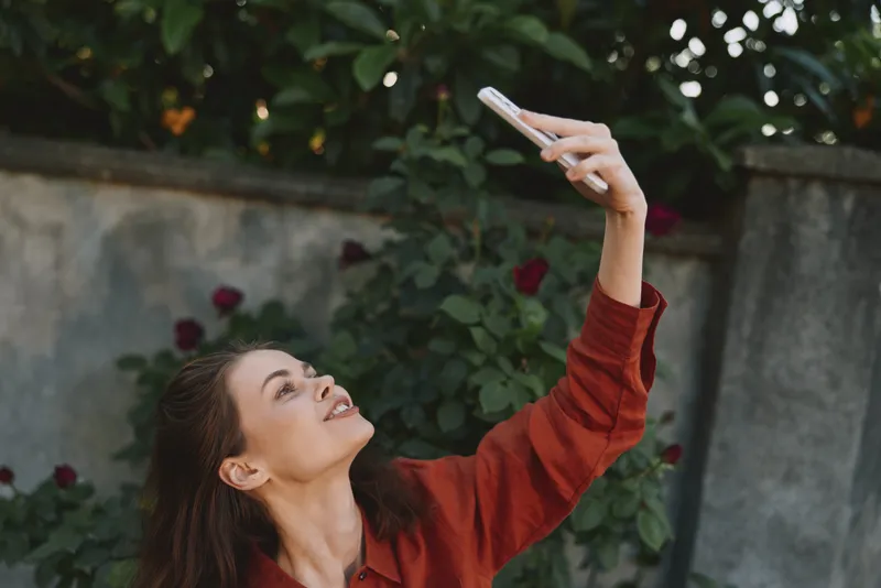 Cheerful woman outdoors, taking a selfie with smartphone, red sweater, warm sunlight, relaxed pose, garden backdrop, casual lifestyle, candid moment