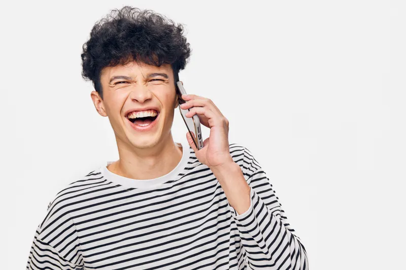 Laughing man talking on mobile phone wearing striped shirt, joyful young adult with curly hair expressing happiness and excitement against white background.