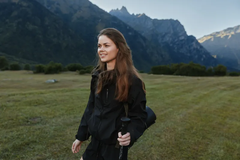 A young woman in outdoor gear stands in a sunlit grassy field with rugged mountains in the distance, smiling softly as she holds a trekking pole and prepares for adventure.