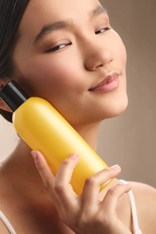asian woman holds a yellow skincare bottle close to her face in a soft, warm setting, exuding calm confidence and a relaxing selfcare moment.