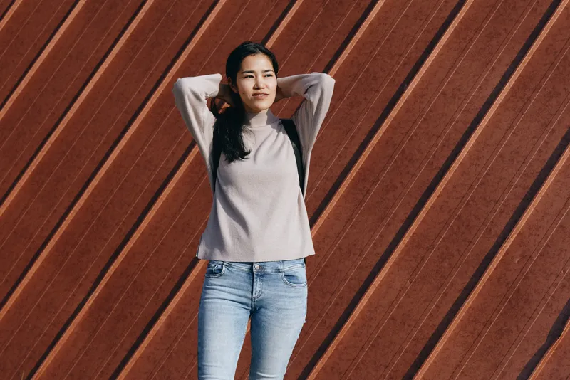 Woman wearing a casual sweater and denim jeans poses against diagonal wooden panels, relaxed stance, outdoor daylight, street style vibe and contemporary fashion sense
