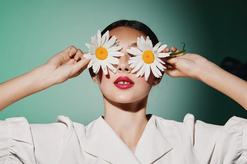 Woman with stylish makeup holding white flowers over her eyes. Artistic beauty and fashion concept in studio with modern green background and soft lighting effect.