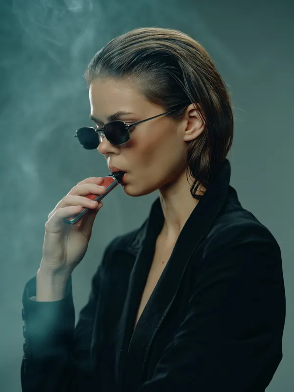 Stylish woman smoking vape with sunglasses and dark outfit in moody atmosphere