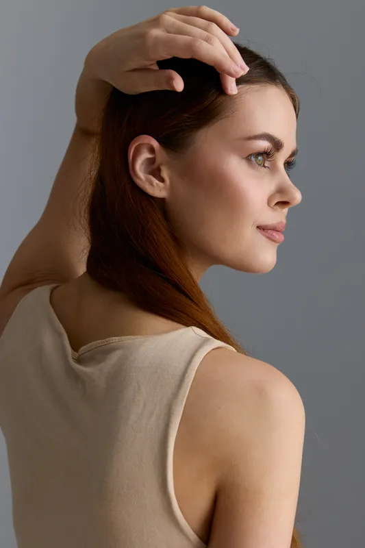 Young woman with long hair posing gracefully in beige sleeveless top against gray background, showcasing natural beauty and calm expression in soft lighting.