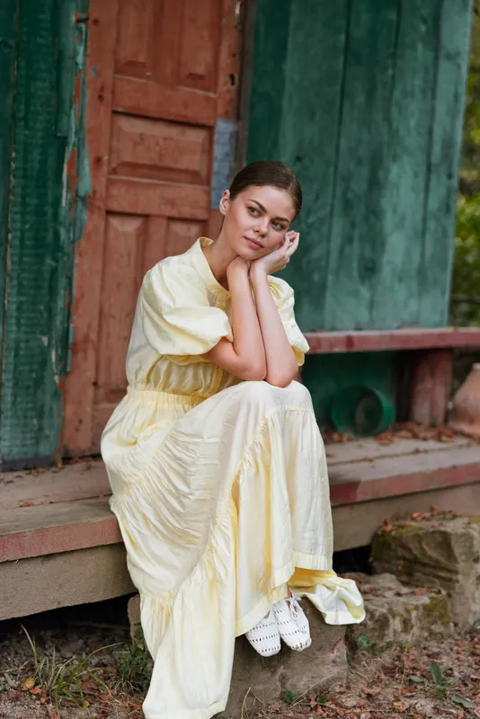 Woman sitting on wooden steps in a pale yellow dress, resting her face on her hands, in a rustic outdoor setting, creating a calm, contemplative portrait with soft natural light.