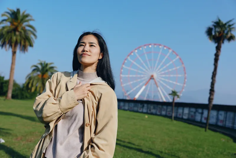Woman in a beige jacket stands outdoors at a sunny park with a large ferris wheel behind, palm trees framing a relaxed, confident pose. A bright day. Sunny.