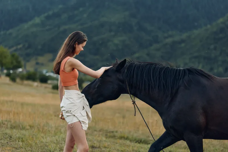 A young woman in casual attire stands in a wide field, gently touching a horse. The scene conveys calm companionship and a serene outdoor moment.