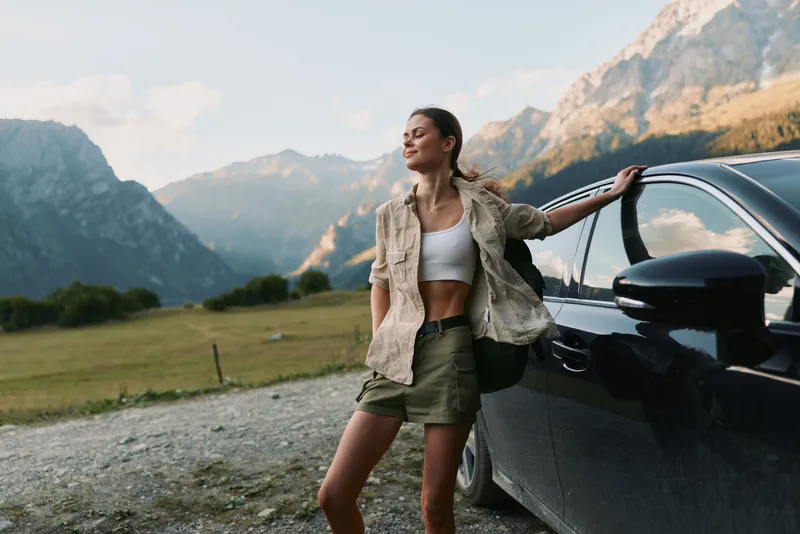 A woman leans against a dark car in a mountain valley, wearing a cropped top and casual jacket, embodying travel, freedom, and outdoor lifestyle in a bright landscape.