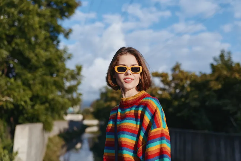 Woman portrait in colorful sweater with sunglasses outdoors, smiling at camera in urban park with trees and blue sky, casual short brown hair and warm sunlight.