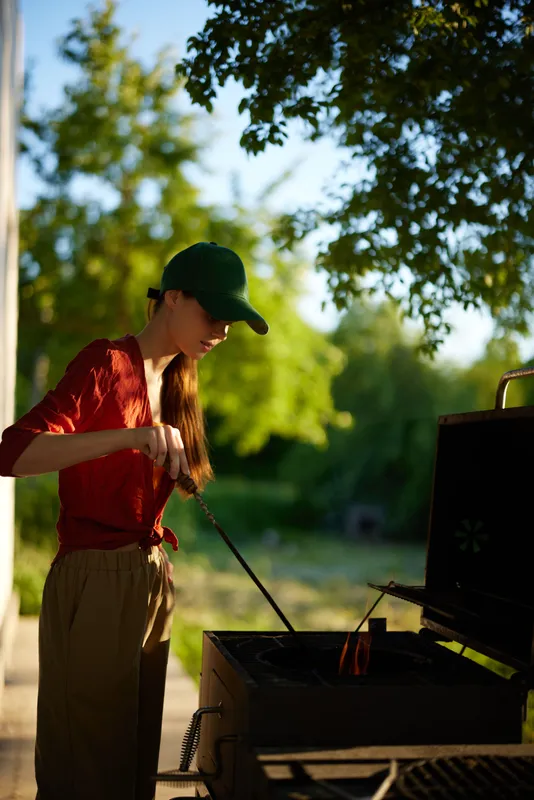 Young woman in casual outfit grilling outdoors in a sunny park setting during summer, expressing focus and enjoyment, surrounded by greenery