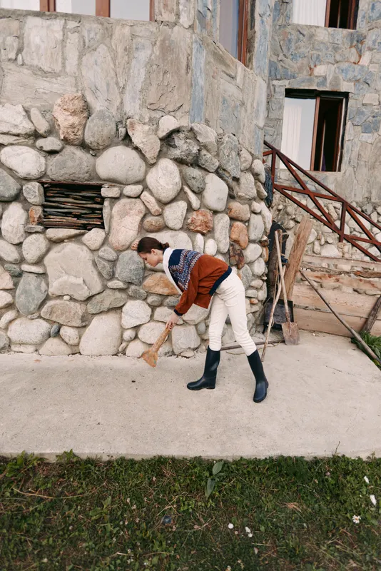 Man sweeping with broom along concrete pathway by stonewall of unfinished building, wearing sweater, white pants and rubber boots, outdoor maintenance and rustic construction chores.