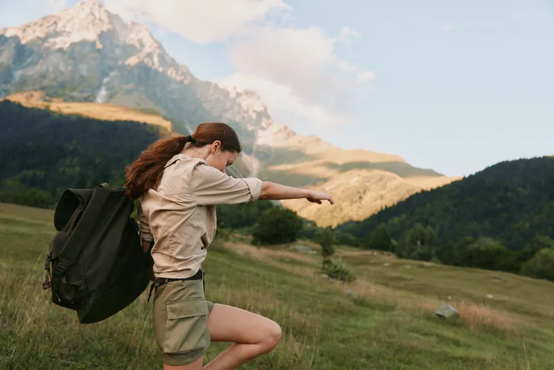 Joyful woman outdoors, mid jump with backpack, sunlit meadow and distant mountains, candid lifestyle moment, vibrant natural colors and active spirit in a bright setting