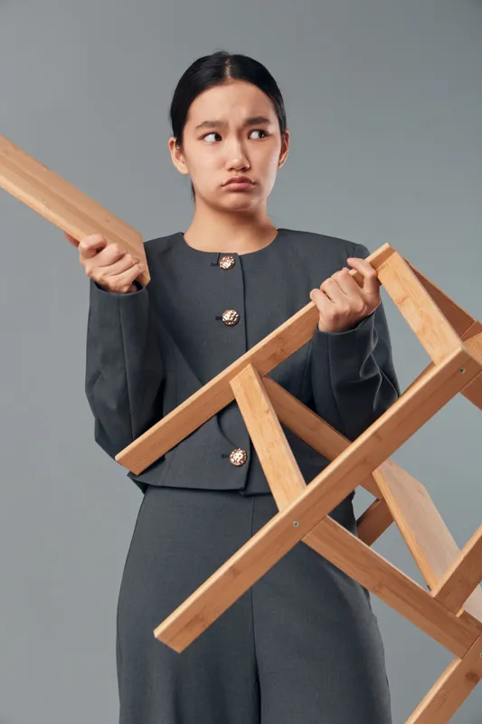 Young woman holding wooden chair with unhappy expression in studio. Female in gray outfit standing with furniture piece showing displeasure and doubt on gray background.