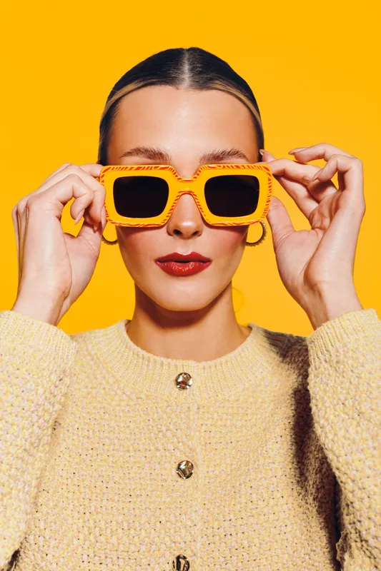 Young woman in yellow sweater adjusts large orange sunglasses on bright yellow background. Fashion portrait with bold colors and confident expression. Concept of style and summer accessories.