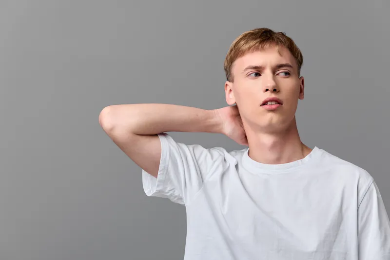 Neck discomfort and strain shown by a man in casual white tshirt stretching his neck in a studio setting with neutral gray background, capturing focused gaze, tense posture, and everyday wellness in